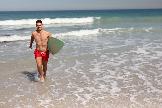 Uomo senza camicia con il surf che cammina sulla spiaggia al sole