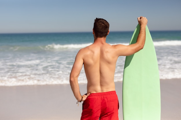 Uomo senza camicia con tavola da surf in piedi sulla spiaggia sotto il sole