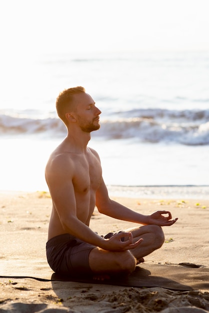 Uomo senza camicia di lato che medita sulla spiaggia