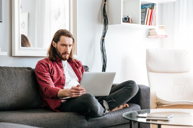 Uomo serio che si siede e che per mezzo del computer portatile sul sofà a casa