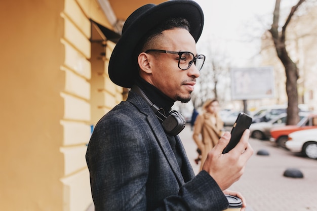 Uomo serio con la pelle marrone che tiene il telefono e guardando la strada. Foto all'aperto del ragazzo africano ben vestito in cappello in attesa amico vicino al caffè.