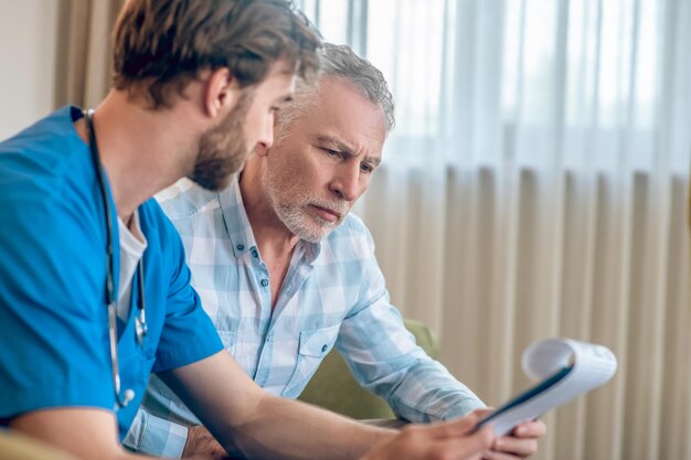 Uomo serio dai capelli grigi focalizzato in una camicia a quadri che guarda un blocco per appunti nelle mani del medico