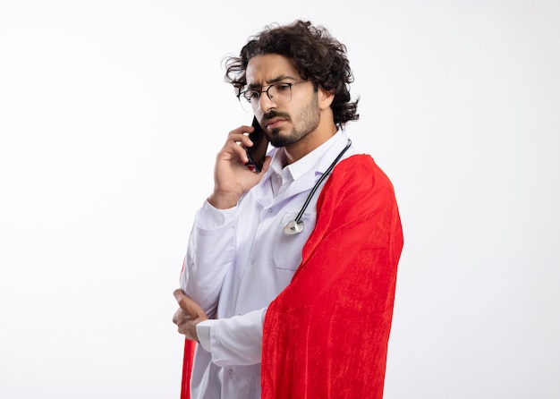 Uomo serio giovane supereroe caucasico in vetri ottici che indossa l'uniforme del medico con mantello rosso e con lo stetoscopio intorno al collo sta lateralmente parlando al telefono con lo spazio della copia