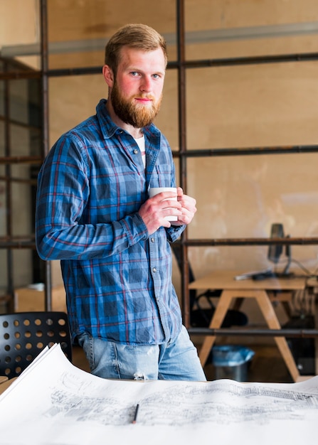 Uomo sicuro che tiene tazza di caffè che sta scrittorio vicino