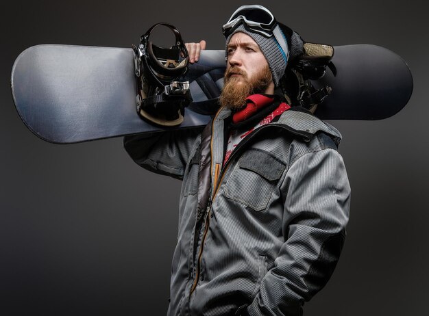 Uomo sicuro con la barba rossa che indossa un'attrezzatura completa che tiene uno snowboard sulla spalla, distogliendo lo sguardo con uno sguardo serio, isolato su uno sfondo scuro.