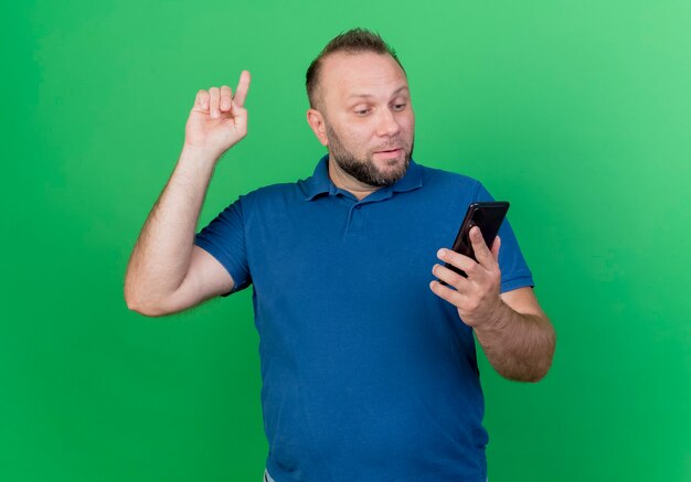 Uomo slavo adulto colpito che tiene e che guarda il telefono cellulare che alza il dito isolato sulla parete verde