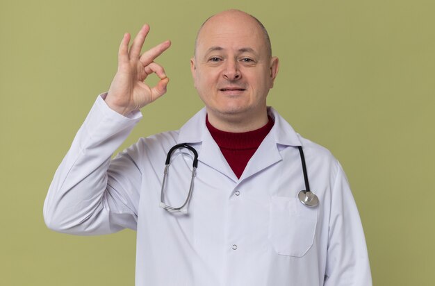 Uomo slavo adulto compiaciuto in uniforme da medico con stetoscopio che gesticola segno ok guardando