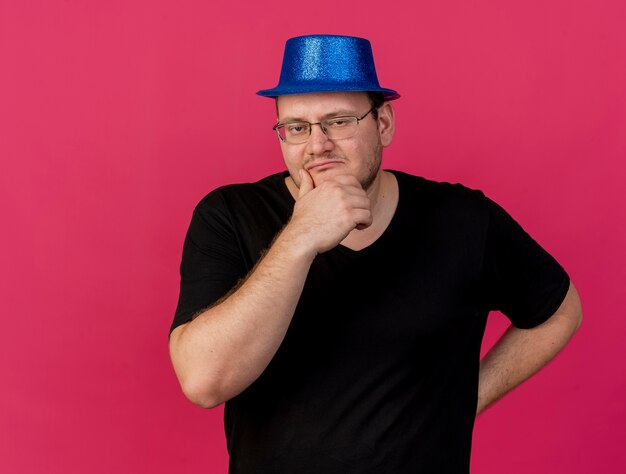 Uomo slavo adulto confuso in occhiali ottici che indossa un cappello da festa blu mette la mano sul mento guardando la telecamera