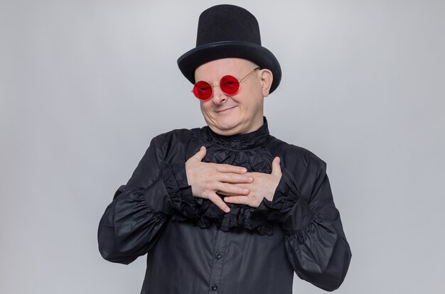 Uomo slavo adulto contento con cappello a cilindro e occhiali da sole in camicia gotica nera che si mette le mani sul petto