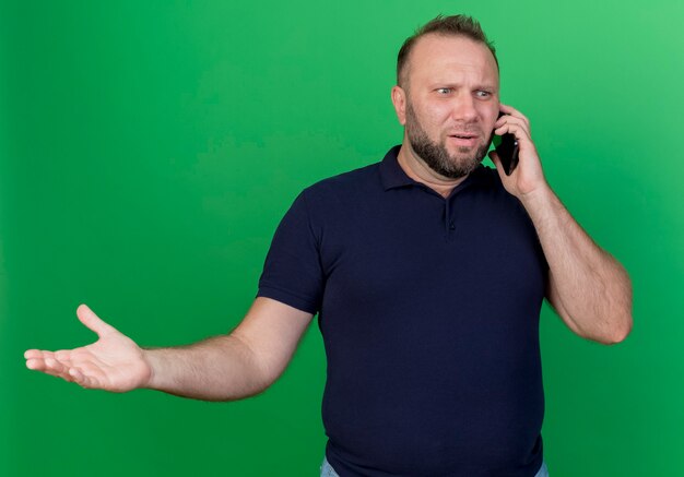 Uomo slavo adulto dispiaciuto parlando al telefono guardando dritto e mostrando la mano vuota isolata sulla parete verde