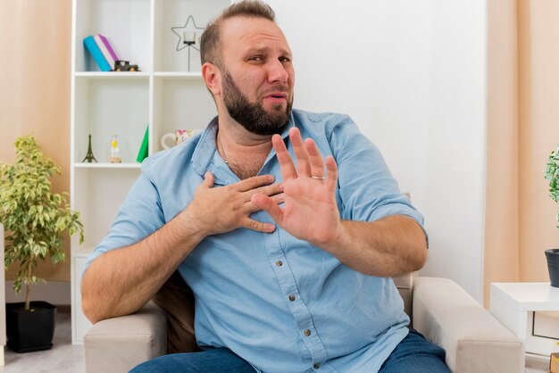 Uomo slavo adulto dispiaciuto si siede sulla poltrona mettendo la mano sul petto e non gesticolando nessun segno di mano all'interno del soggiorno