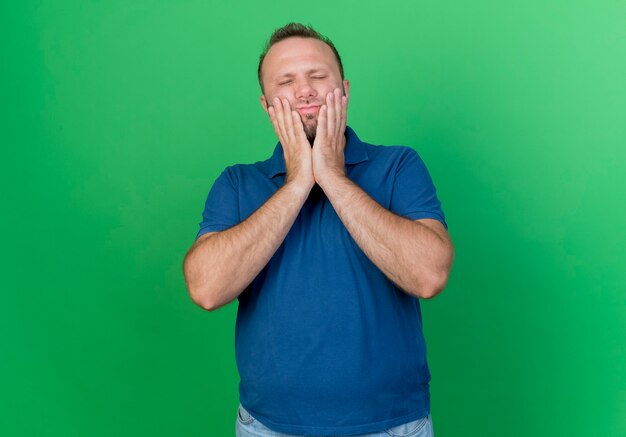 Uomo slavo adulto dolorante che mette le mani sul viso che soffre di mal di denti con gli occhi chiusi isolato sulla parete verde con lo spazio della copia