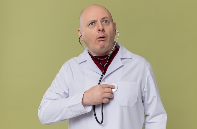 Uomo slavo adulto impressionato in uniforme da medico che ascolta il suo battito cardiaco con lo stetoscopio