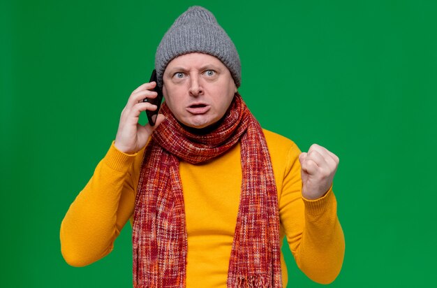 Uomo slavo adulto infastidito con cappello invernale e sciarpa intorno al collo che parla al telefono e tiene il pugno
