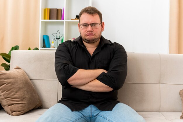 Uomo slavo adulto offeso in vetri ottici si siede sulla poltrona con le braccia incrociate all'interno del soggiorno