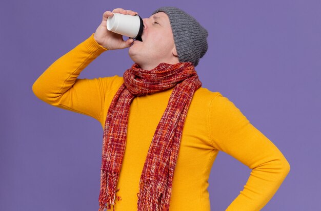 Uomo slavo adulto scontento con cappello invernale e sciarpa intorno al collo che beve da un bicchiere di carta