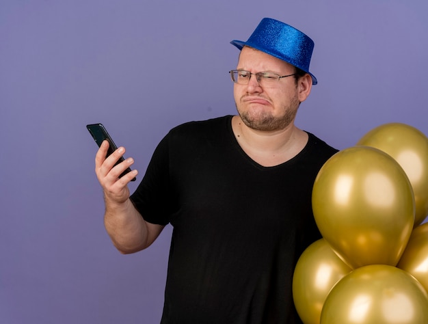 Uomo slavo adulto scontento in occhiali ottici che indossa un cappello da festa blu tiene palloncini di elio e telefono and