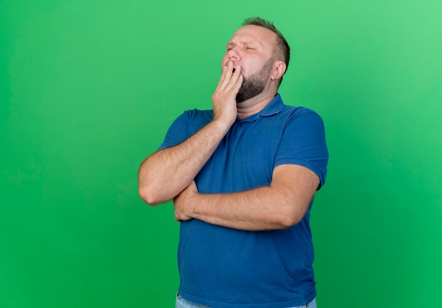 Uomo slavo adulto stanco che mette la mano sulla bocca che sbadiglia con gli occhi chiusi isolati sulla parete verde con lo spazio della copia