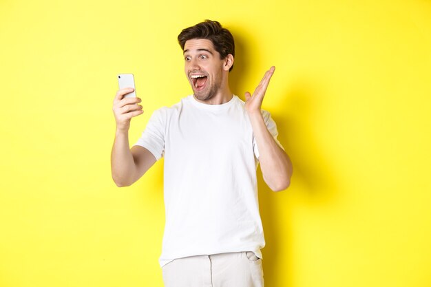 Uomo sorpreso e felice che guarda lo schermo del telefono cellulare, legge notizie fantastiche, in piedi su sfondo giallo