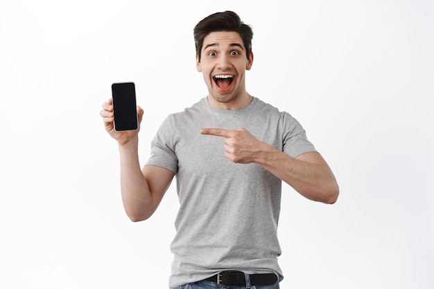 Uomo sorpreso e stupito che punta allo schermo vuoto dello smartphone che mostra l'applicazione del negozio online di visualizzazione dell'applicazione in piedi su sfondo bianco