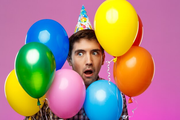 Uomo sorpreso festeggia il compleanno, tenendo baloons colorati sul muro viola.