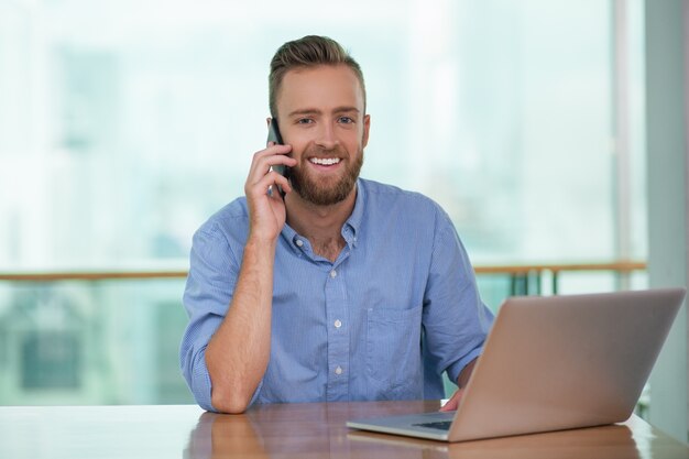 Uomo sorridente che chiama il telefono e di lavoro sul computer portatile