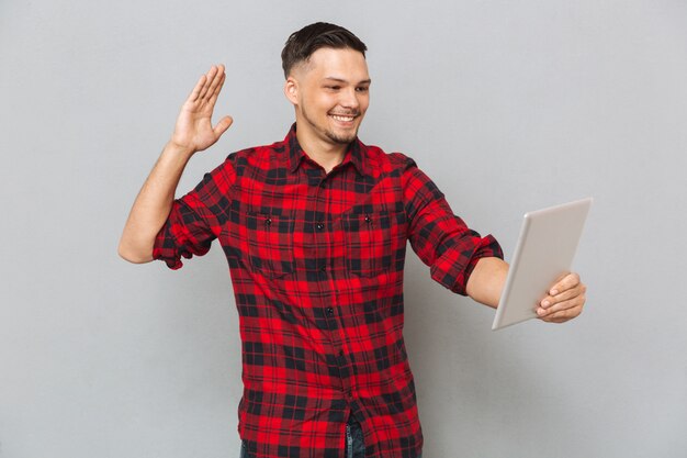 Uomo sorridente che per mezzo del computer della compressa e ondeggiando alla macchina fotografica