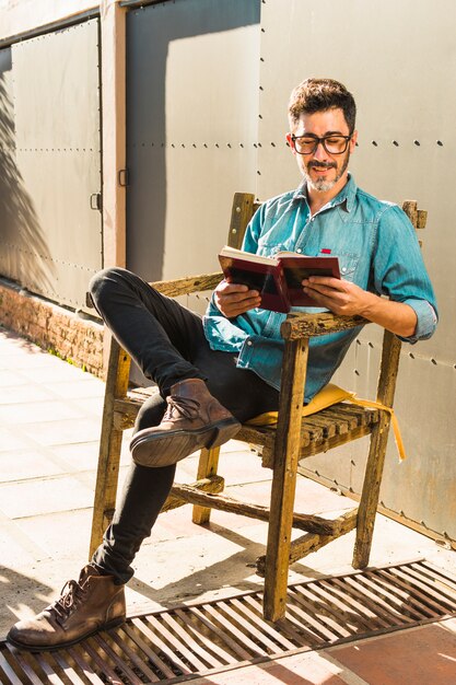 Uomo sorridente che si siede sulla sedia di legno leggendo il libro