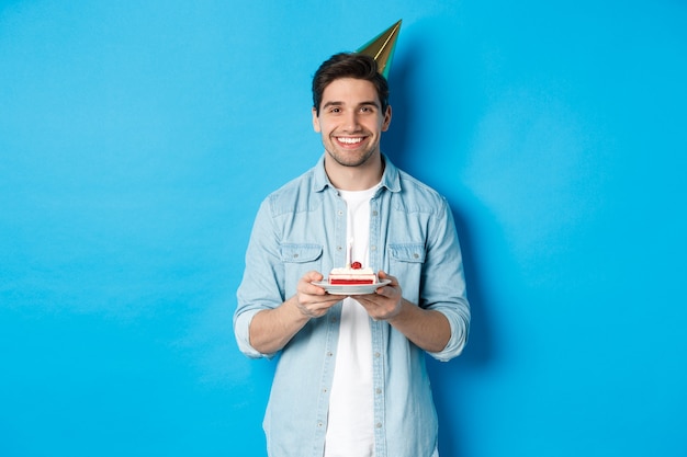 Uomo sorridente che tiene la torta di b-day e indossa un cappello da festa di compleanno, celebrando su sfondo blu.