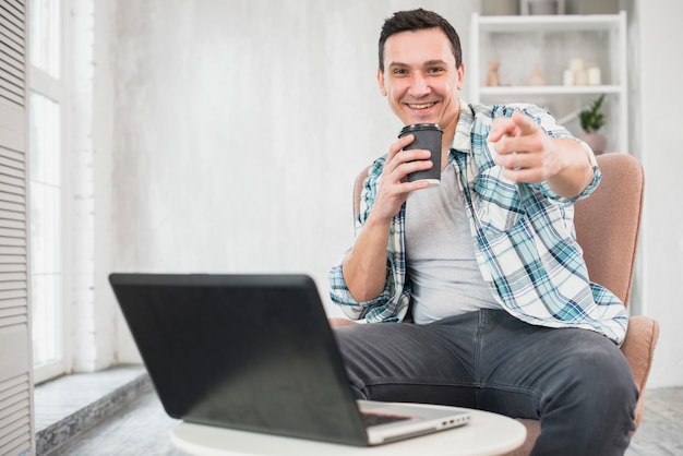 Uomo sorridente che tiene tazza della bevanda sulla sedia vicino al computer portatile a casa