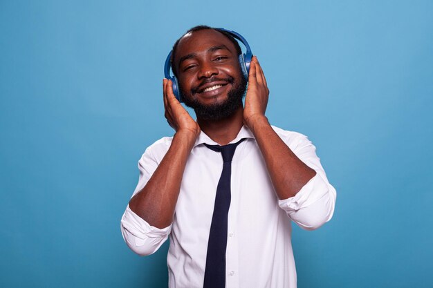 Uomo sorridente che tocca le cuffie wireless sentendosi rilassato godendosi una piacevole musica di successo su sfondo blu. Rilassante uomo d'affari in camicia bianca podcast in streaming audio in cuffia auricolare.