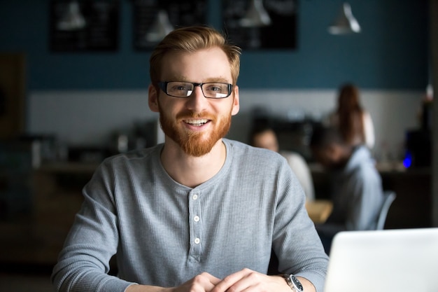 Uomo sorridente della testarossa con il computer portatile che esamina macchina fotografica in caffè