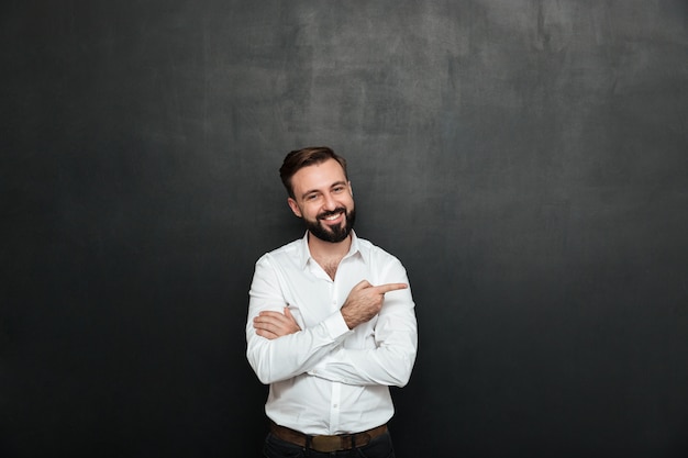 Uomo sorridente in camicia bianca che posa sulla macchina fotografica con il vasto sorriso, indicante il dito indice da parte sopra lo spazio grigio scuro della copia