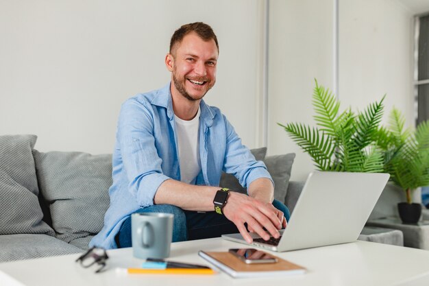 uomo sorridente in camicia seduto rilassato sul divano a casa a tavola lavorando online su laptop da casa