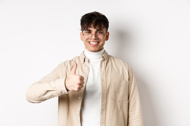 Uomo sorridente positivo che mostra i pollici in su e sembra soddisfatto, raccomandando e approvando la cosa, in piedi soddisfatto su sfondo bianco