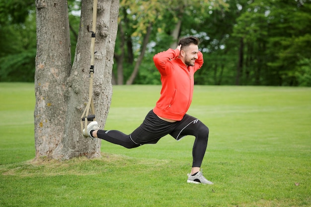 Uomo sportivo che si esercita con l'imbracatura dell'istruttore di sospensione in città Parco sotto gli alberi estivi per il fitness sportivo Uomo felice in giacca rossa