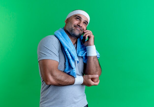 Uomo sportivo maturo in fascia con un asciugamano intorno al collo parlando al telefono cellulare con il sorriso sul viso in piedi sopra la parete verde