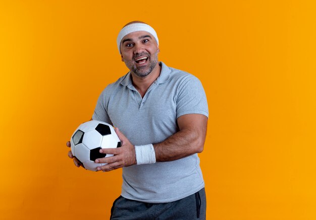 Uomo sportivo maturo in fascia tenendo il pallone da calcio guardando in avanti con il sorriso sul viso in piedi sopra il muro arancione 2