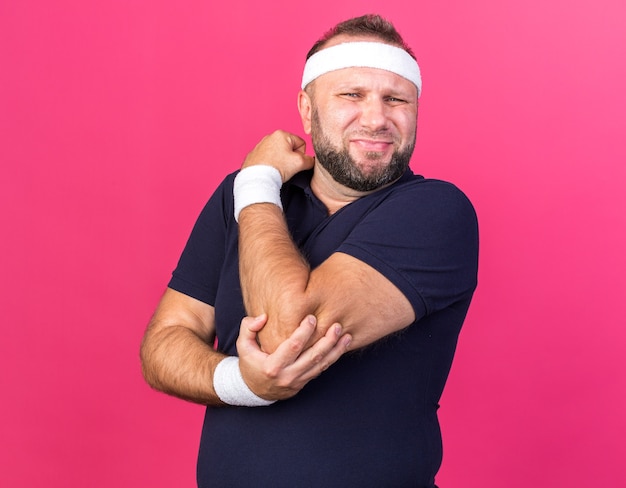 uomo sportivo slavo adulto dolorante che indossa fascia e braccialetti che tengono il gomito isolato sul muro rosa con spazio di copia