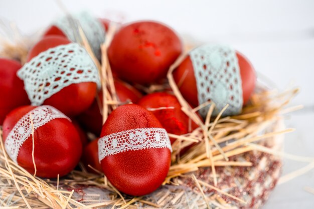 Uova di Pasqua rosse in un nido di fieno, legate un nastro di pizzo, primo piano, sdraiato su un legno bianco