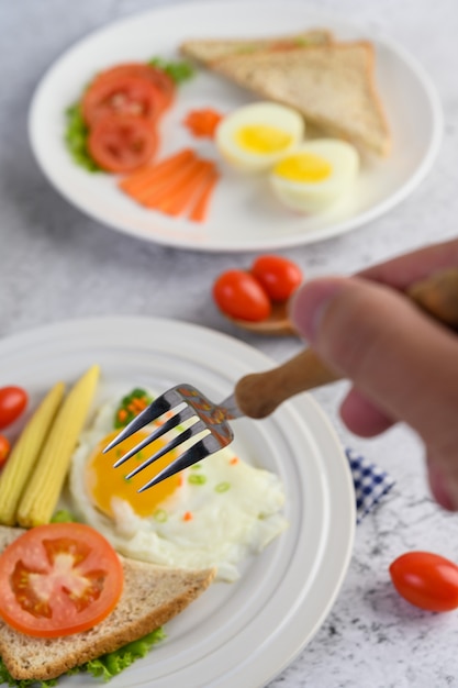 Uova fritte, pane, carote e pomodori su un piatto bianco per la colazione, messa a fuoco selettiva palmare con una forchetta.