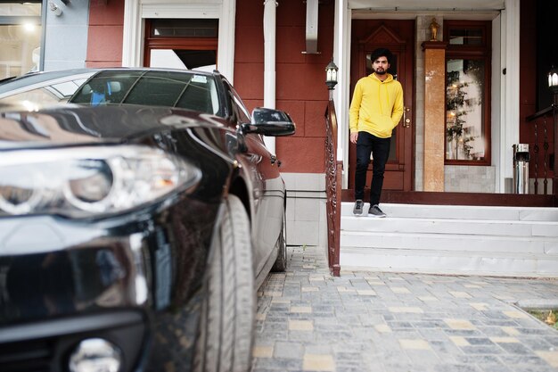 Urban giovane hipster indiano uomo in una felpa gialla alla moda Cool sud asiatico ragazzo indossare felpa con cappuccio stare sul portico della casa con macchina nera d'affari