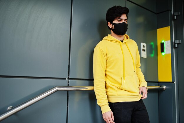 Urban giovane uomo indiano hipster in una felpa gialla alla moda stare vicino all'ingresso del codice Cool sud asiatico ragazzo indossare felpa con cappuccio e maschera nera per la protezione del viso durante la nuova normalità