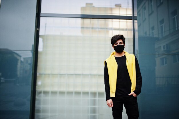 Urbano giovane hipster indiano in una felpa gialla alla moda Il ragazzo del sud asiatico indossa una felpa con cappuccio e una maschera nera per proteggere il viso durante la nuova normalità