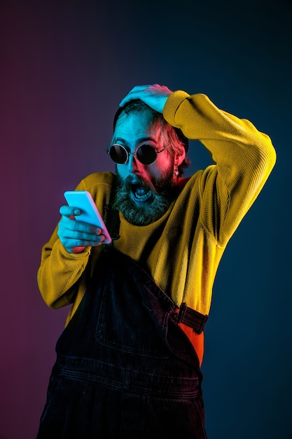 Usando il telefono, estremamente scioccato. Ritratto dell'uomo caucasico sul fondo dello studio sfumato in luce al neon. Bellissimo modello maschile con stile hipster. Concetto di emozioni umane, espressione facciale, vendite, annuncio.