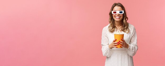 Uscire per il tempo libero e concetto di primavera Ritratto di felice ragazza bionda spensierata in abito bianco godendo di guardare film in 3d indossando occhiali mangiare popcorn e sorridere frequentare cinema sfondo rosa