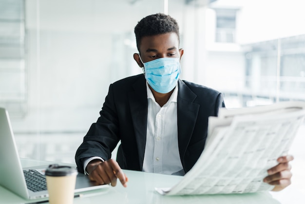 Usura africana bella dell'uomo d'affari nella maschera medica con il giornale di mattina vicino all'ufficio del centro di affari