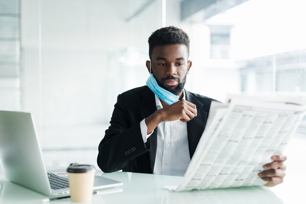 Usura africana bella dell'uomo d'affari nella maschera medica con il giornale di mattina vicino all'ufficio del centro di affari