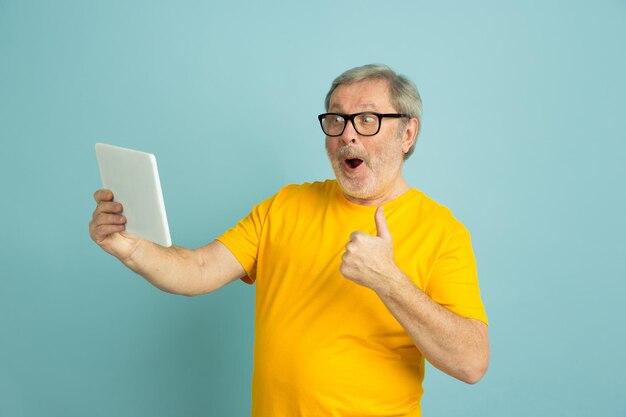 Utilizzando tablet, mostrando il pollice in su. Ritratto di uomo caucasico isolato su sfondo blu studio. Bello modello maschio in camicia gialla.