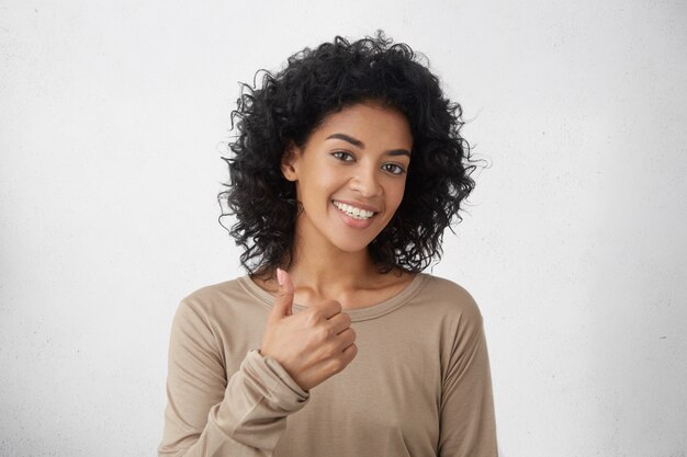 Va molto meglio. Bella giovane donna entusiasta allegra della corsa mista con capelli ricci che mostrano i pollici sul gesto